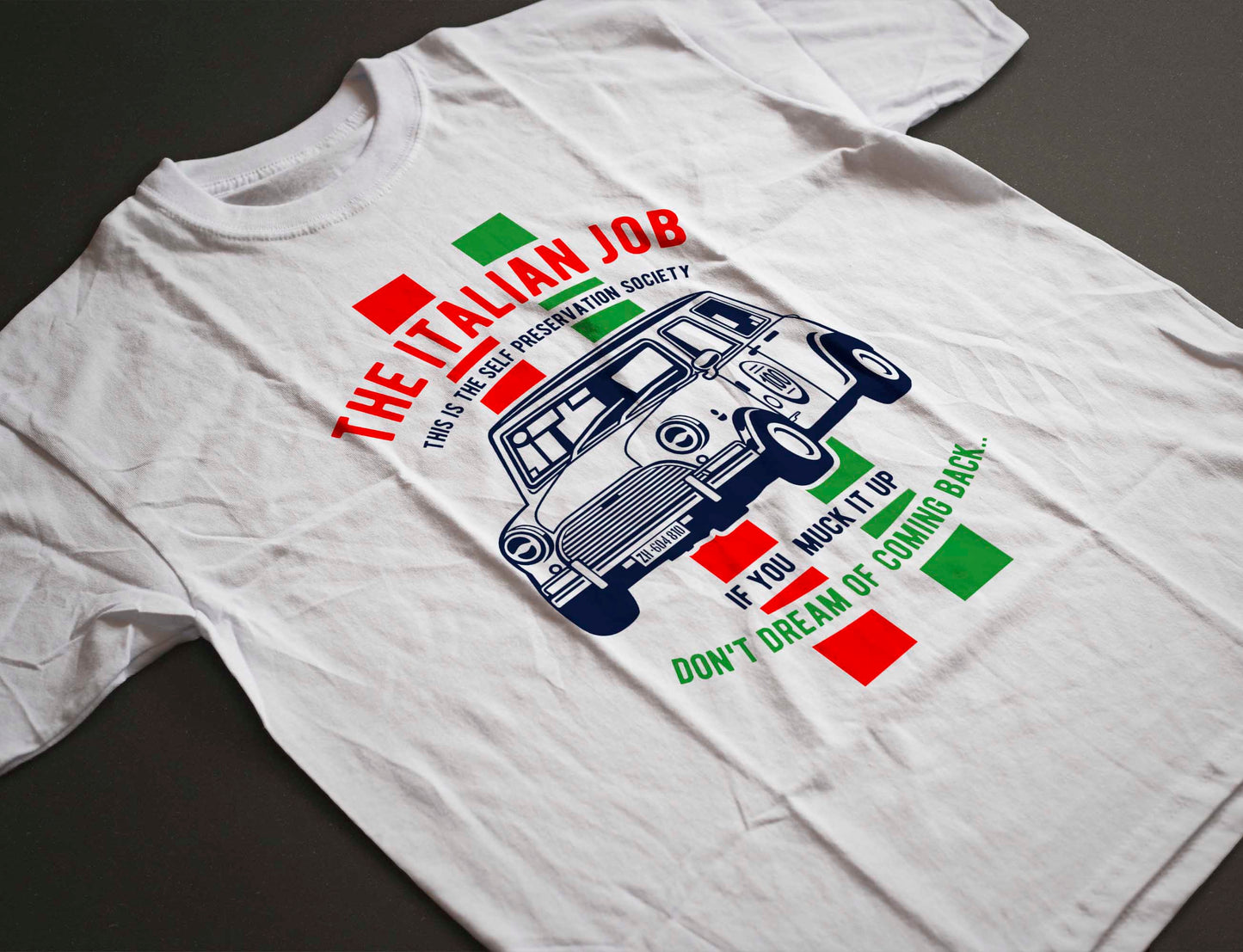 the white t-shirt displays a bold design with a blue mini cooper car in the center. the text "the italian job" arches above the car, and below, the warning "if you muck it up, don't dream of coming back". the phrase "this is the self preservation society" is also part of the design, complemented by red and green blocks on either side of the car.