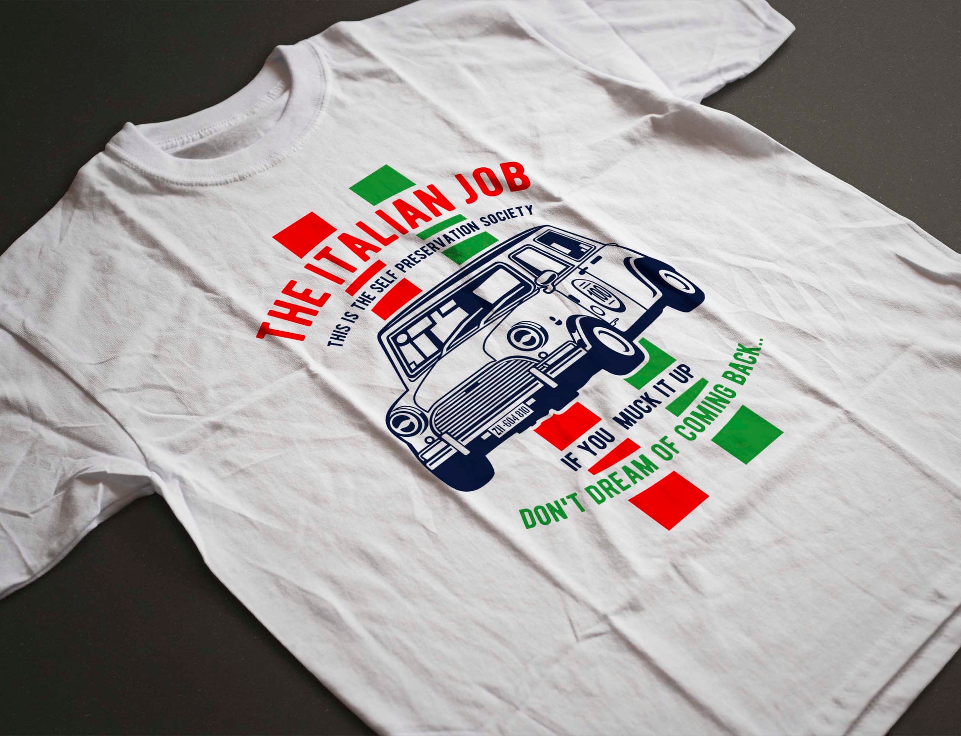 The white t-shirt displays a bold design with a blue Mini Cooper car in the center. The text "The Italian Job" arches above the car, and below, the warning "If you muck it up, don't dream of coming back". The phrase "This is the Self Preservation Society" is also part of the design, complemented by red and green blocks on either side of the car.