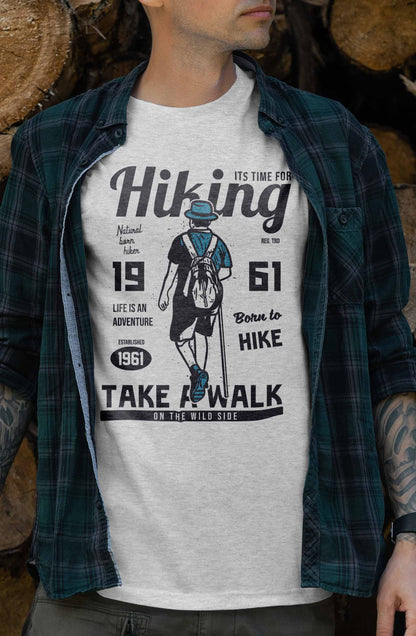 The t-shirt artwork includes a stylized depiction of a hiker ready for an adventure, complemented by various phrases such as "Natural born hiker," "Life is an adventure," and "Born to Hike 1961". The overall theme celebrates hiking and exploration, encouraging wearers to embrace their wild side.