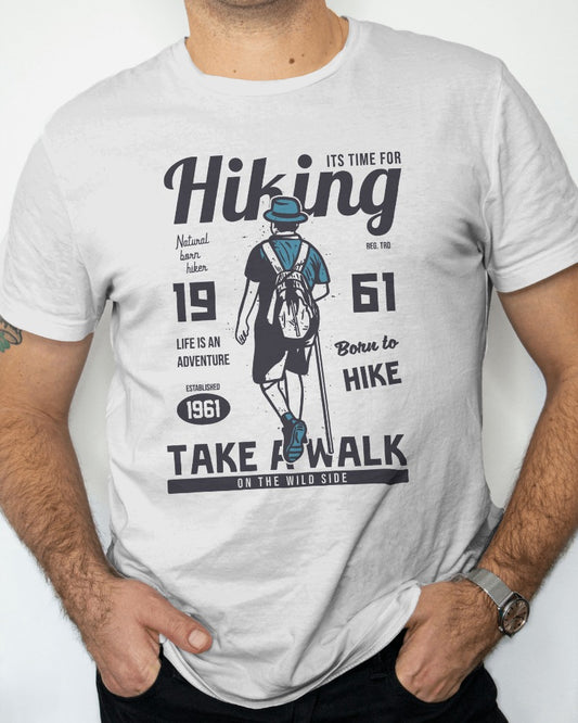 This design on the t-shirt shows a hiker from behind, dressed in hiking gear with a backpack, walking towards a destination. The text "Hiking - It's Time For," "Born to Hike," and "Natural born hiker" in white and blue colors add to the adventurous theme, emphasizing a life of outdoor exploration since 1961.