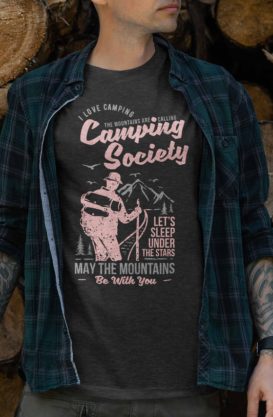 This t-shirt design depicts a hiker in silhouette, facing a mountain landscape. Above and below the hiker are phrases like "I Love Camping, The Mountains Are Calling," and "Let's Sleep Under the Stars, May the Mountains Be With You," all in stylized pink and white fonts on a dark background.
