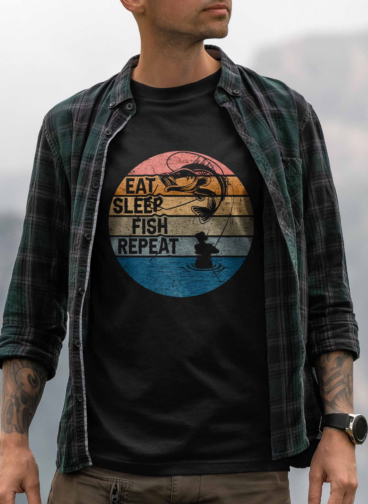 a t-shirt design showcases a stylized circular emblem divided into colorful segments, representing different times of the day, with "eat sleep fish repeat" written in bold. the top depicts a fish jumping out of the water against a sunset, and the bottom shows the calm silhouette of a fisherman fishing from his boat.