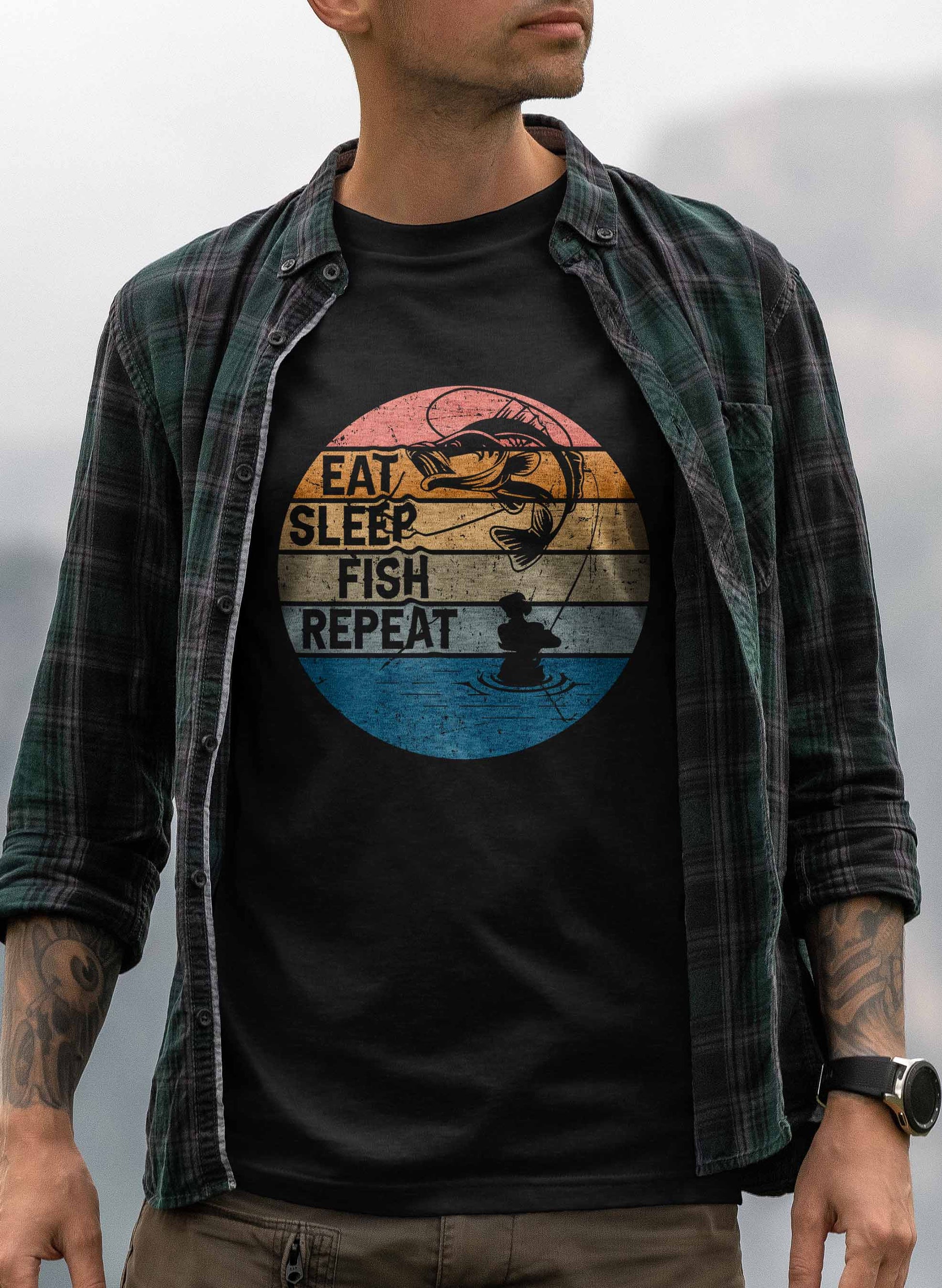 A t-shirt design showcases a stylized circular emblem divided into colorful segments, representing different times of the day, with "Eat Sleep Fish Repeat" written in bold. The top depicts a fish jumping out of the water against a sunset, and the bottom shows the calm silhouette of a fisherman fishing from his boat.