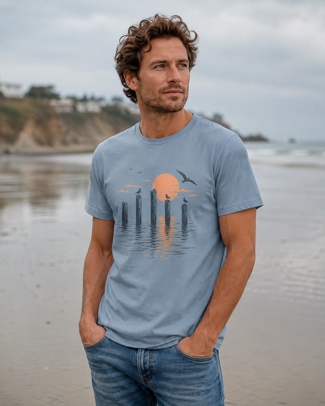 Man wearing a stone blue graphic T-shirt featuring seabirds perched on wooden pier posts beneath an orange coastal sunset, photographed on a beach.