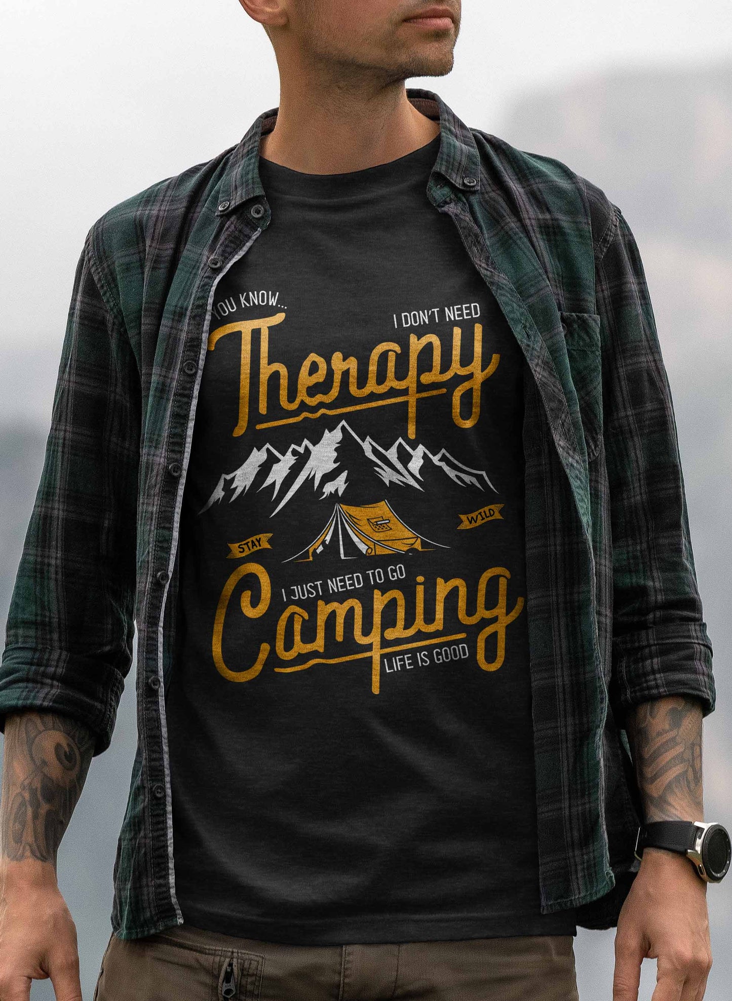 the image features a black t-shirt with a graphic that reads "therapy - i don't need therapy, i just need to go camping - life is good". the design includes stylized mountains in the background with a tent in the foreground, all in white and yellow on a black background.